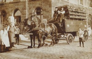 Historische Pferdekutsche im Q21 - „Historische Fotografie eines von zwei Pferden gezogenen Bierwagens vor einem Backsteingebäude, beladen mit Bierflaschen und umgeben von mehreren Männern in Arbeitskleidung.“